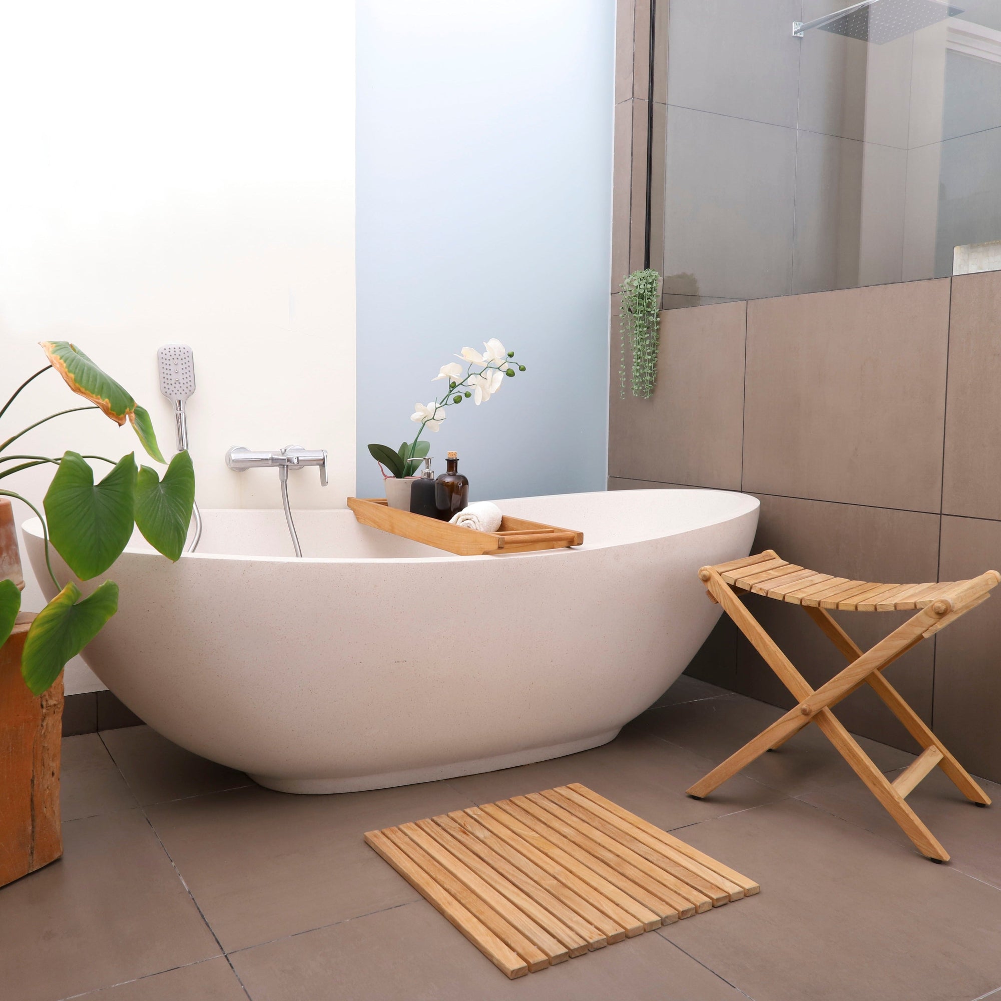 A teak wood roll-up shower mat with rubber footing placed on a tiled bathroom floor, next to a bathtub and a wooden stool, with a natural finish.